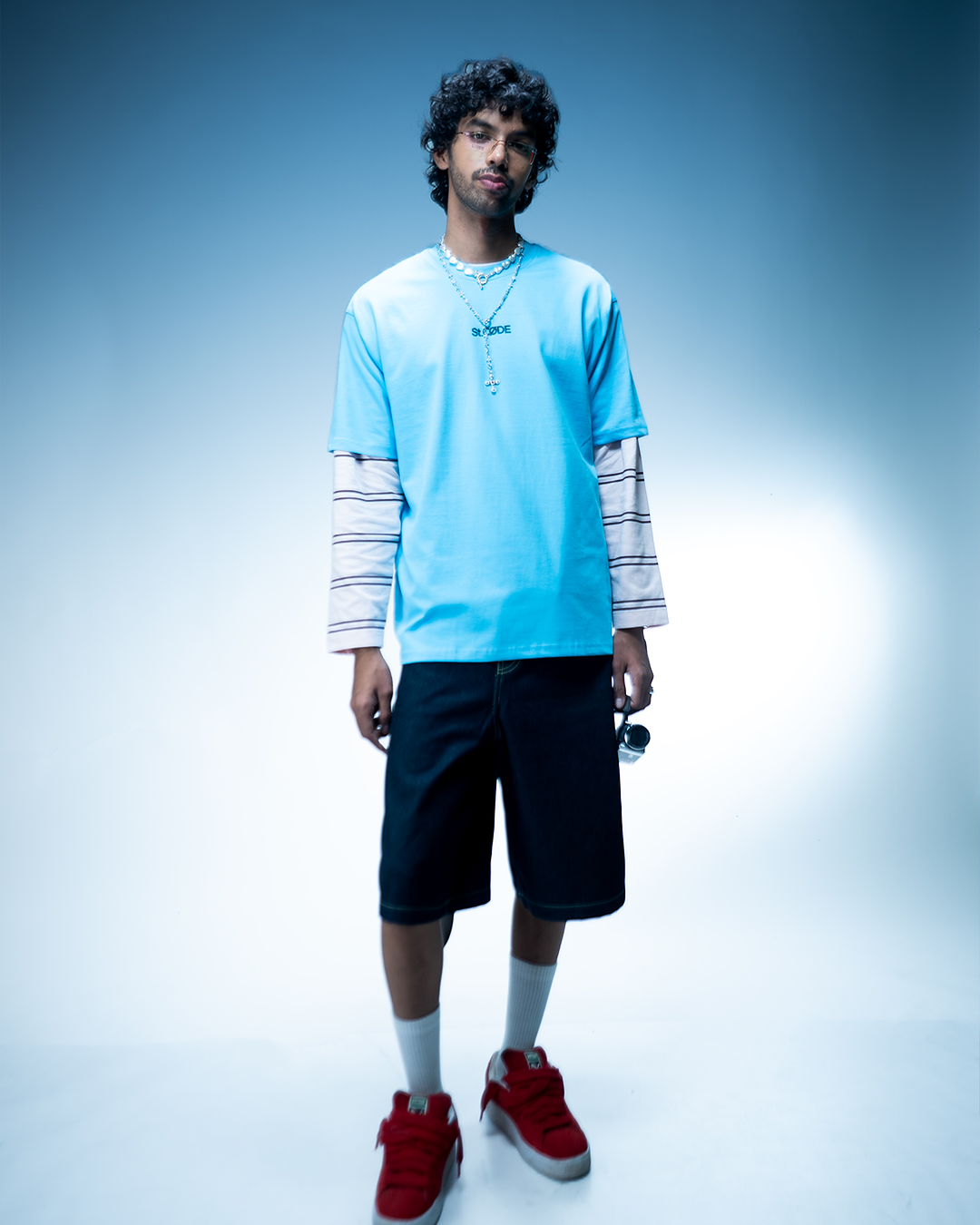 Person wearing a light blue shirt with a logo, black shorts, red shoes, and white socks on a gradient background.